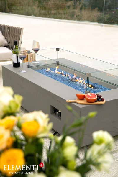 Moderner rechteckiger Gaskamin-Tisch von Elementi mit blauen Glassteinen, Flammen und Glas-Windschutz auf einer hellen Terrasse. Elementi Logo.