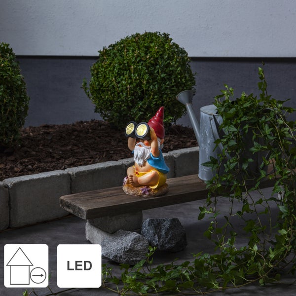 Dekorativer Gartenzwerg mit Fernglas und LED-Beleuchtung auf einer Gartenbank neben einer Gießkanne und Pflanzen.