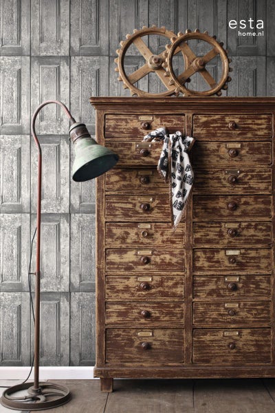 Dekorative Szene mit Holzkommode, Zahnrädern und Stehlampe vor einer Tapete mit Holzpaneel-Optik.