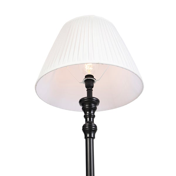 Stehlampe mit schwarzem, verziertem Metallgestell und weißem, plissiertem Lampenschirm.