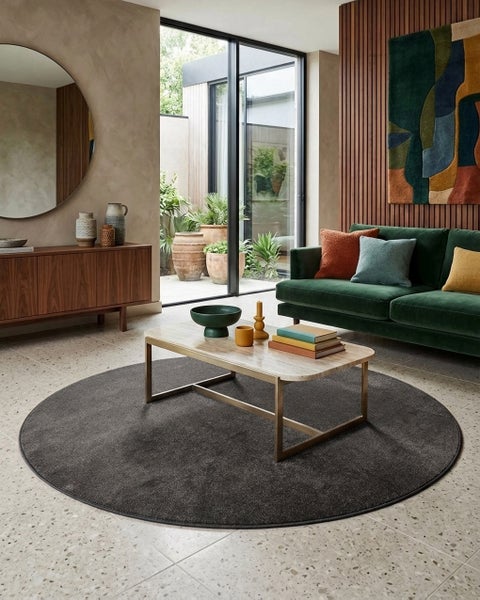 Moderner Wohnbereich mit rundem dunkelgrauem Teppich, grünem Samtsofa, Couchtisch mit Marmorplatte und Holzwandpaneelen vor Fensterfront.