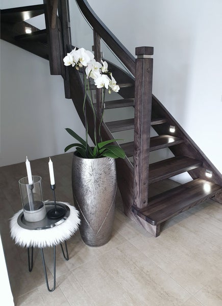 Dunkle Holztreppe mit Glasgeländer und LED-Beleuchtung, hohe strukturierte Metallvase mit Orchideen und Beistelltisch in hellem, modernem Flur.