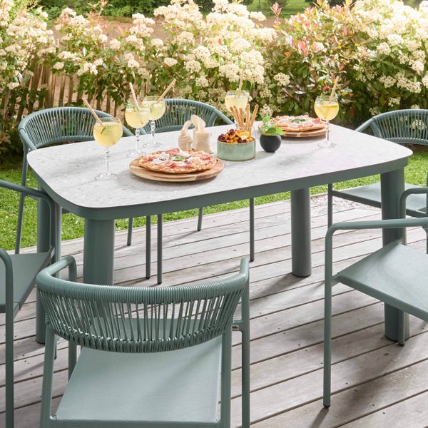 Modernes, helles Gartenmöbel-Set mit einem rechteckigen Tisch in Steinoptik und Stühlen mit Seilgeflecht auf einer Holzterrasse.