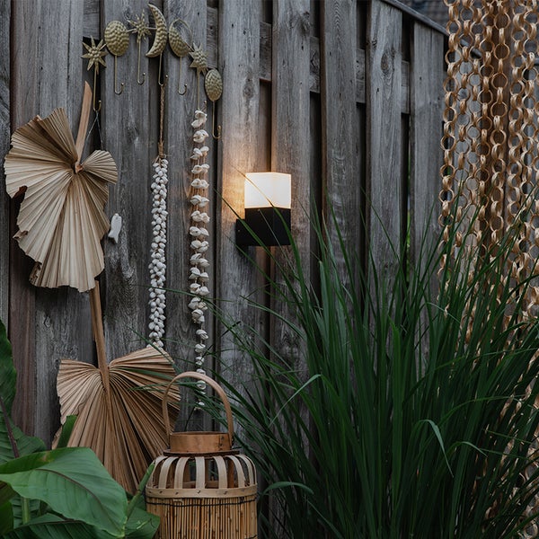 Eckige Außenwandleuchte mit schwarzer Halterung und warmem Licht an einem Holzzaun mit getrockneten Palmblättern und Muschelgirlanden.