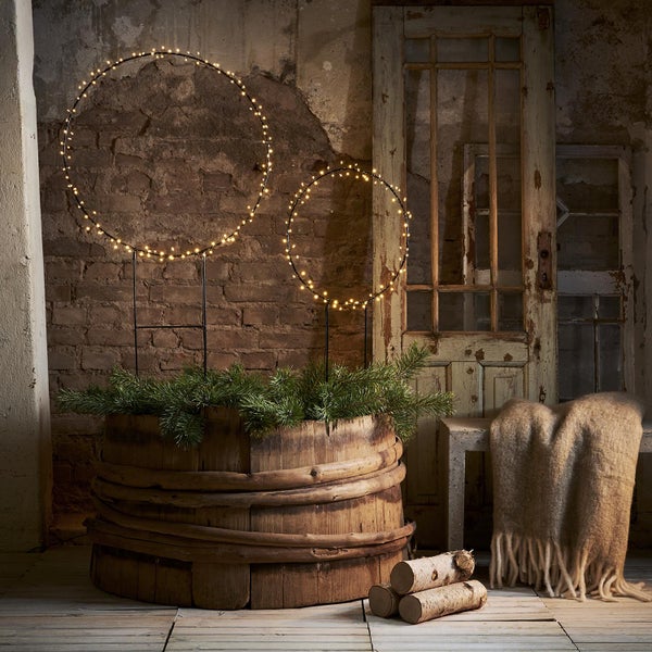 Dekorative Szene mit zwei beleuchteten Metallringen, Holzbottich mit Tannenzweigen, Holzscheiten und Wolldecke