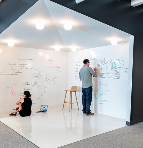 Heller, hausförmiger Bürobereich mit beschreibbaren weißen Wänden, runden Deckenleuchten und Personen, die an Skizzen arbeiten.