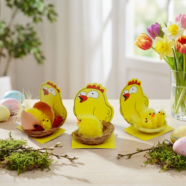 Osterdekoration mit drei stehenden Pappaufstellern in Kükenform, kleinen Stoffküken in Nestern und frühlingshafter Tischdeko mit Blumen und Eiern.
