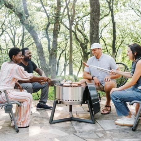 Vier Personen sitzen auf einer hellen Steinterrasse im Garten um einen runden silbernen Holzkohlegrill und bereiten gemeinsam Würstchen zu.