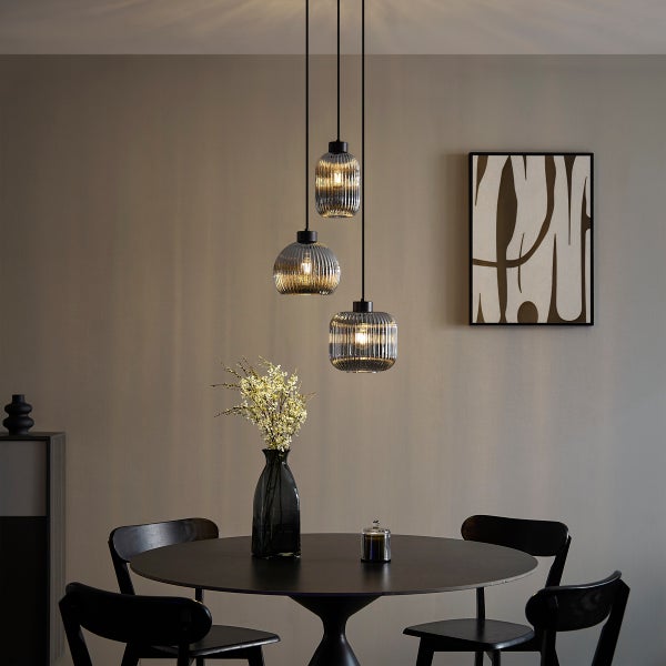 Moderne dreiflammige Pendelleuchte mit geriffelten Glasschirmen über einem dunklen runden Esstisch in einem minimalistischen Raum.