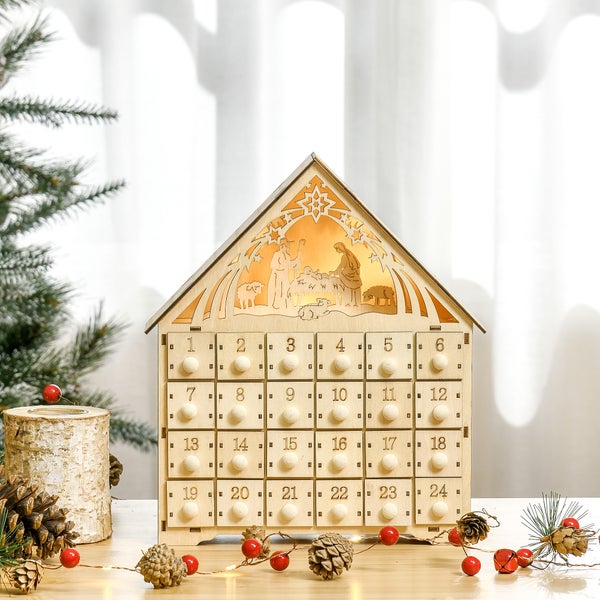 Adventskalender aus Holz in Hausform mit 24 Schubladen und beleuchteter Krippenszene, umgeben von Tannenzweigen, Zapfen und roten Beeren.