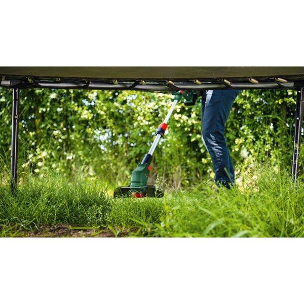 Eine Person trimmt mit einem Akku Rasentrimmer das Gras unter einem Trampolin im Garten.