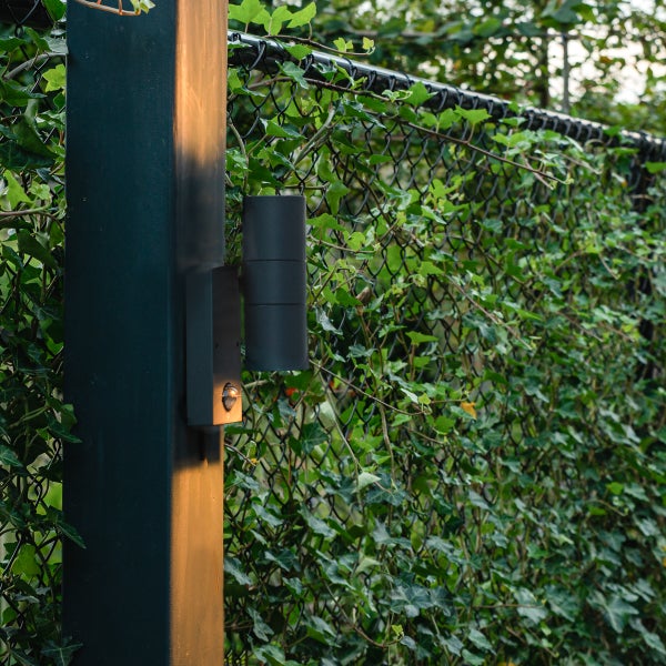 Moderne Außenwandleuchte mit Bewegungsmelder, montiert an einem Pfosten vor einem mit Efeu bewachsenen Zaun.