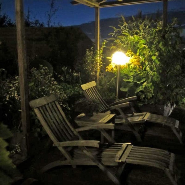 Zwei Garten-Liegestühle aus Holz mit Fußstützen und Beistelltisch unter einer Pergola bei Nacht, beleuchtet durch eine warme Gartenleuchte.