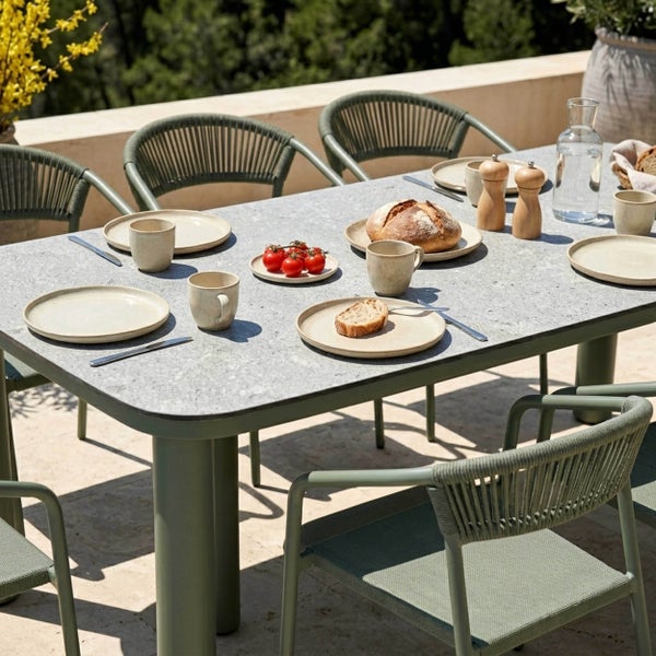 Moderne Garten-Essgruppe auf einer Terrasse. Rechteckiger Tisch in Steinoptik mit abgerundeten Ecken und grünen Stühlen mit Seilgeflecht-Rückenlehne.