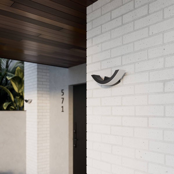 Moderne, gebogene Außenwandleuchte an einer weißen Ziegelwand im Eingangsbereich mit Holzdecke und Pflanzen.