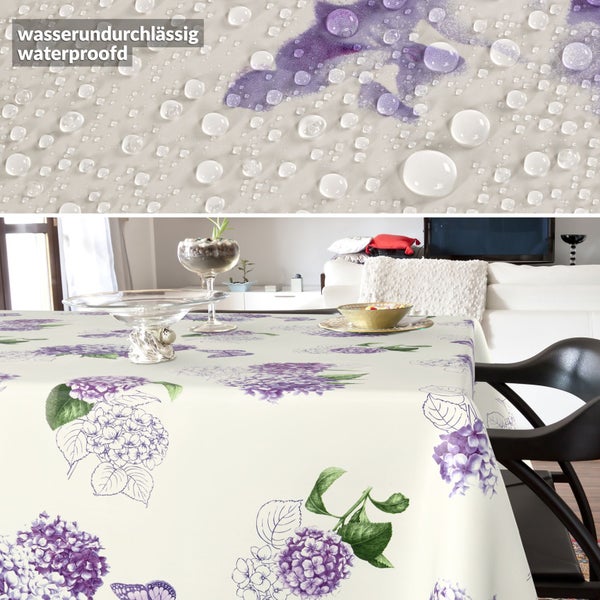 Wasserabweisende Tischdecke mit lila Hortensien-Muster in einem hellen Wohnzimmer. Nahaufnahme zeigt Wassertropfen auf dem Stoff.