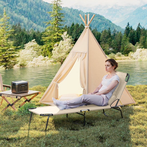 Frau auf einer klappbaren Campingliege neben einem Tipi-Zelt und einem hölzernen Klapptisch mit tragbarer Powerstation in Ufernähe.