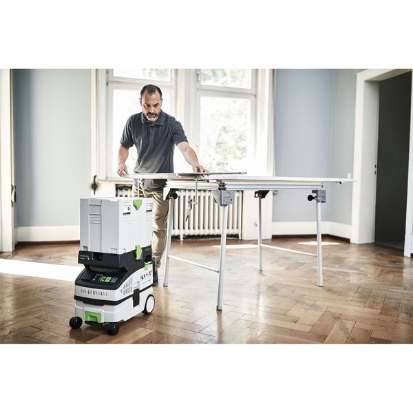 Festool Absaugmobil und Multifunktionstisch in einem hellen Raum mit Parkettboden, ein Mann arbeitet am Tisch.