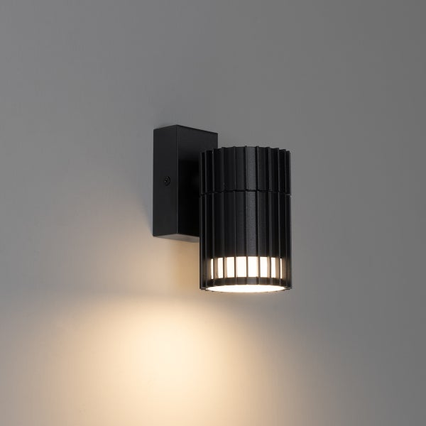 Schwarze zylindrische Außenwandleuchte mit vertikalen Rillen und warmweißem Lichtschein nach unten.