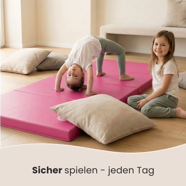 Zwei Kinder spielen auf einer pinkfarbenen Turnmatte mit Kissen im Zimmer.