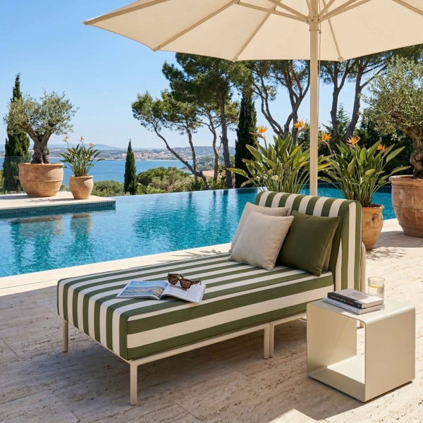 Gestreifte Sonnenliege mit Kissen am Pool unter einem Sonnenschirm. Beistelltisch, Pflanzen und Meerblick. Helle, organisierte Gartenlandschaft.