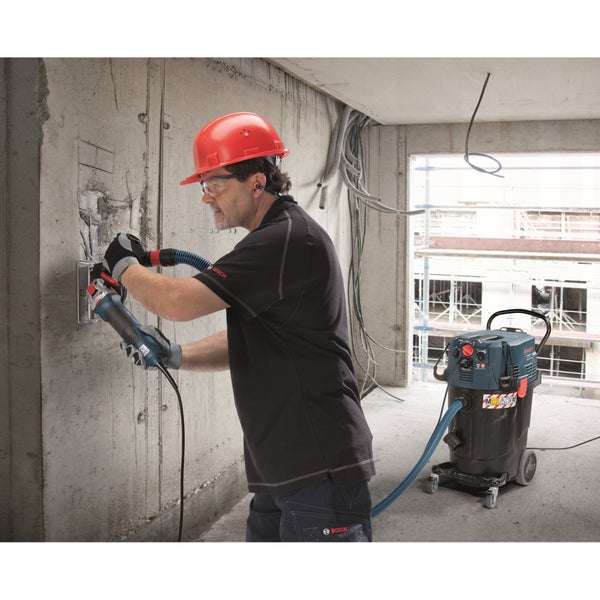 Handwerker mit Bosch Winkelschleifer und angeschlossenem Industriesauger bei der Arbeit an einer Betonwand auf einer Baustelle.