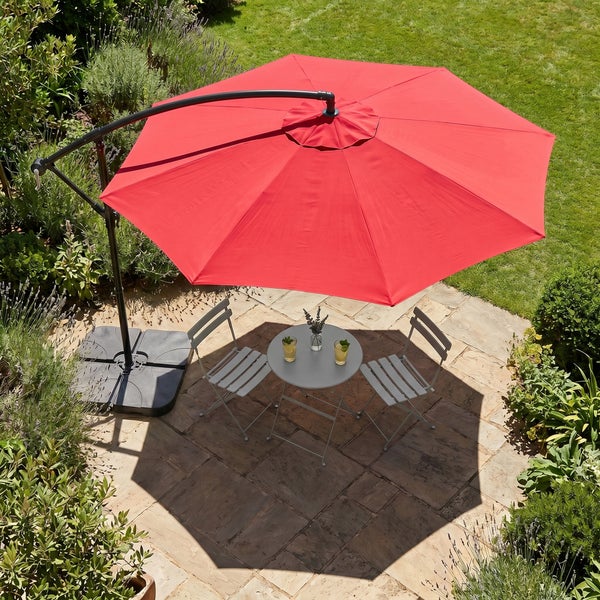 Großer roter Ampelschirm über einem Bistro-Set mit Tisch und zwei Stühlen auf einer Steinterrasse in einem Garten.