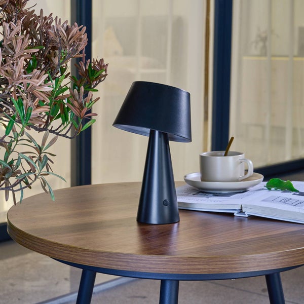 Moderne schwarze Tischleuchte auf einem runden Holztisch neben einer Kaffeetasse und einer Zeitschrift.