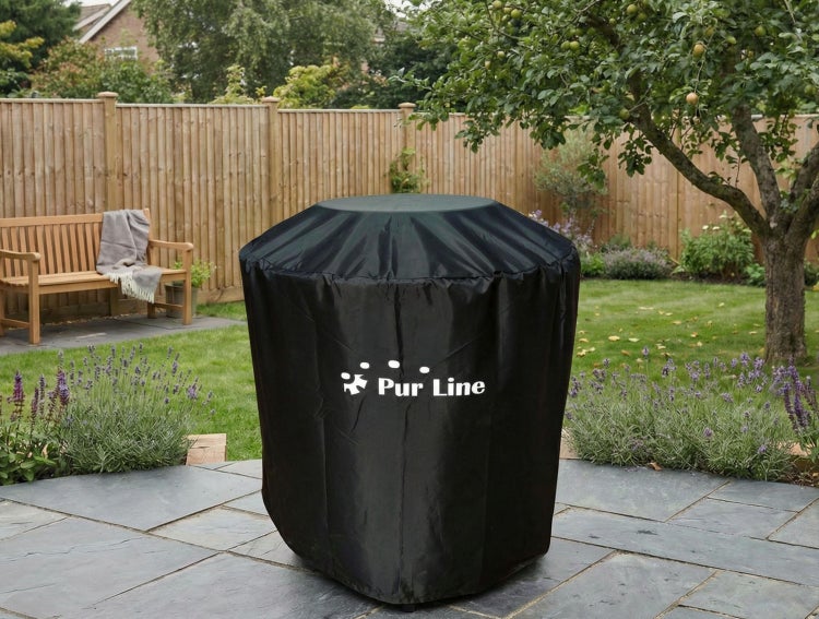 Schwarze Schutzhülle mit Pur Line Logo auf einer Steinterrasse in einem hellen, organisierten Garten mit Rasen, Holzzaun und Holzbank.
