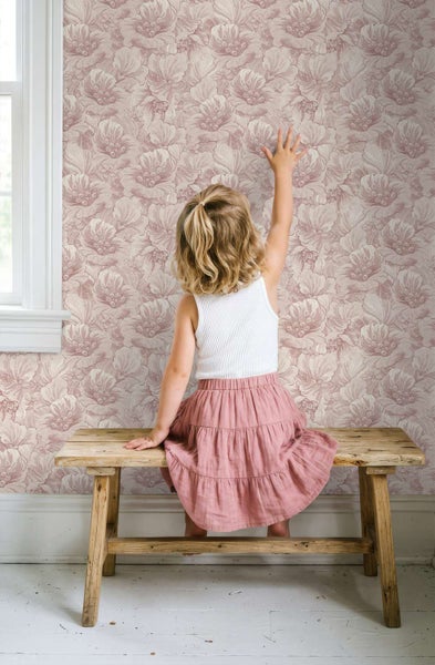 Ein kleines Mädchen sitzt auf einer rustikalen Holzbank vor einer Wand mit floraler Mustertapete und streckt die Hand aus.