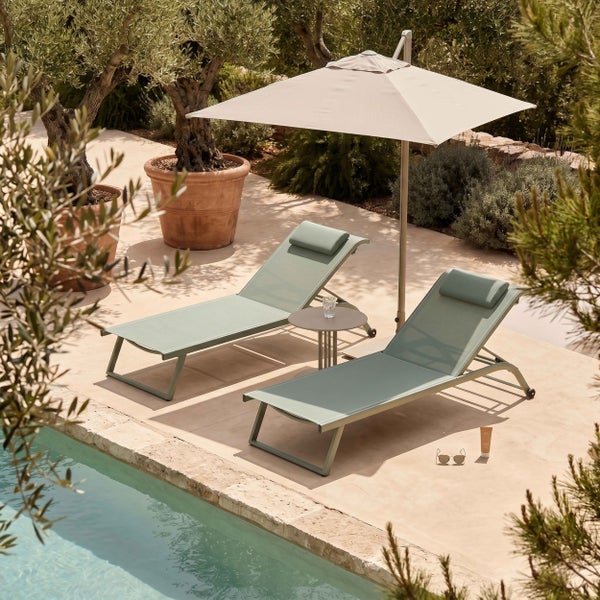 Zwei Gartenliegen mit Kopfstützen, Beistelltisch und Sonnenschirm auf einer Steinterrasse am Pool zwischen Olivenbäumen.