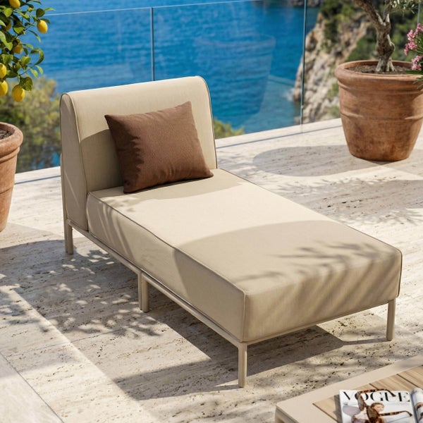 Moderne Gartenliege mit beigem Polster und braunem Kissen auf einer hellen Terrasse mit Meerblick.