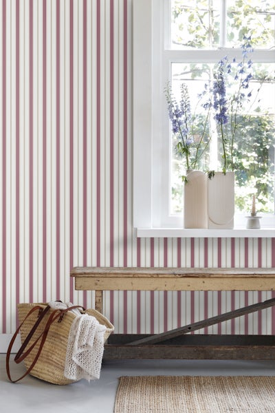 Helle Wand mit gestreifter Tapete hinter einer Holzbank, Fensterbank mit Blumenvasen, geflochtener Tasche und Sisalteppich.