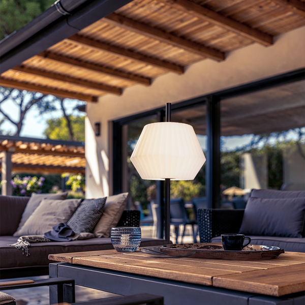 Moderne Außen-Tischleuchte mit weißem Schirm auf einem Holztisch im Terrassenbereich mit Lounge-Möbeln unter einer Holzpergola.