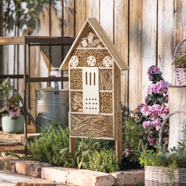 Insektenhotel aus Holz mit verschiedenen Füllmaterialien wie Bambus und Stroh in einem Garten mit Pflanzen und Blumen.