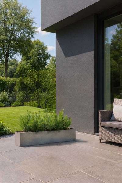 Rechteckiger Pflanzkasten in Steinoptik mit Pflanzen auf einer modernen Terrasse aus Steinplatten neben einer verputzten Hauswand.