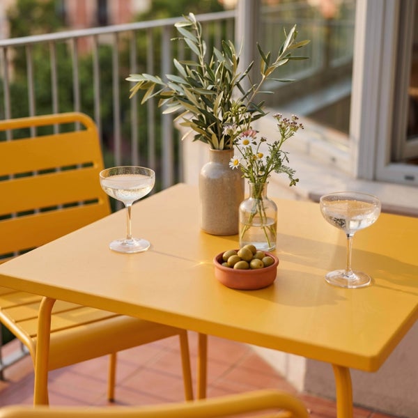 Gelbes Bistro-Set aus Metall auf einem Balkon mit quadratischem Tisch, passenden Stühlen, Gläsern, Oliven und Blumenvasen.