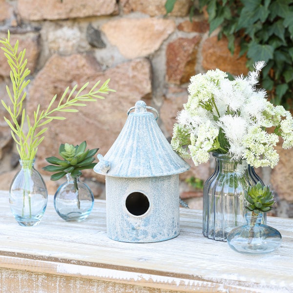 Rustikales Vogelhaus aus Metall in Hellblau neben Glasvasen mit Kunstpflanzen und weißen Blumen auf hellem Holztisch vor einer Steinwand.