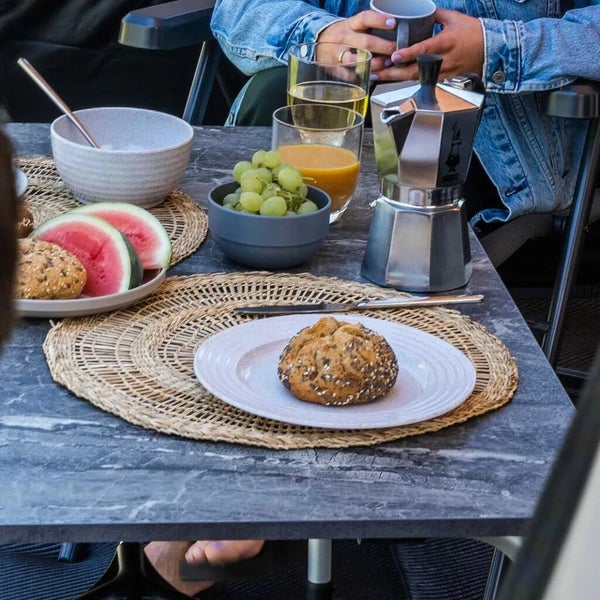 Gedeckter Gartentisch in Marmoroptik mit Espressokocher, Brötchen, Obst und Gläsern auf runden Tischsets aus Naturgeflecht.