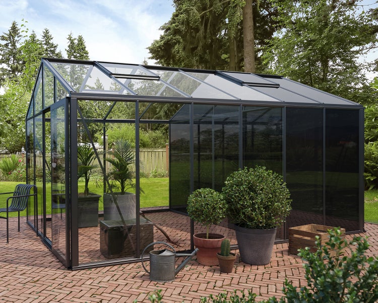 Modernes Gewächshaus aus Glas mit schwarzem Aluminiumrahmen und Dachfenstern auf einer gepflasterten Terrasse im hellen Garten.