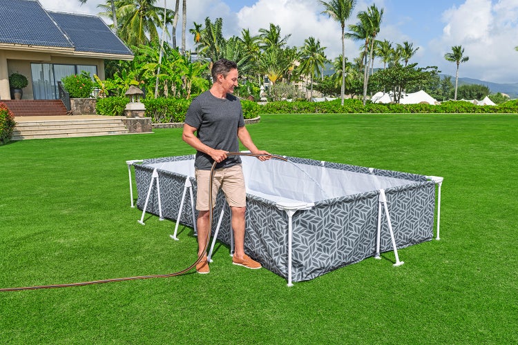 Ein Mann füllt einen rechteckigen Rahmenpool mit Blattmuster auf einer grünen Rasenfläche mit einem Gartenschlauch mit Wasser.