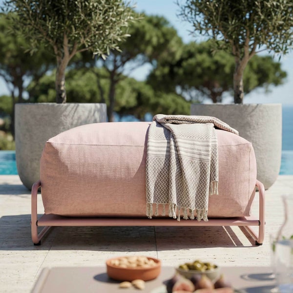 Moderner Outdoor-Hocker mit rosa Stoffbezug auf Metallgestell, mit Decke auf einer sonnigen Terrasse neben Olivenbäumen.