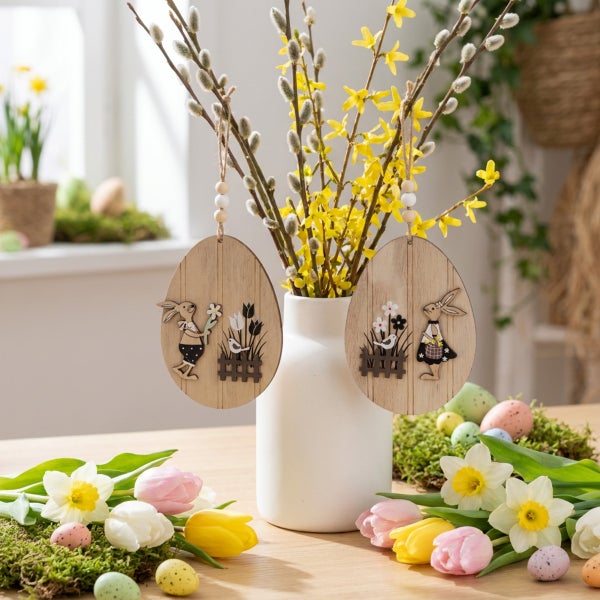 Zwei hängende Ostereier aus Holz als Dekoration an einer Vase mit Weidenkätzchen und Forsythienzweigen auf einem Tisch mit Blumen und Eiern.