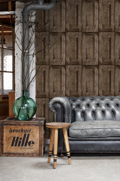 Raumansicht mit Vintage-Tapete, Ledersofa, Holzhocker und dekorativer Glasvase auf einer Holzkiste