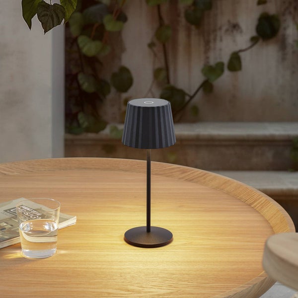 Helle, geordnete Außenszene mit schwarzer kabelloser Tischleuchte auf einem runden Holztisch neben einem Wasserglas und Magazin vor Pflanzenwand.