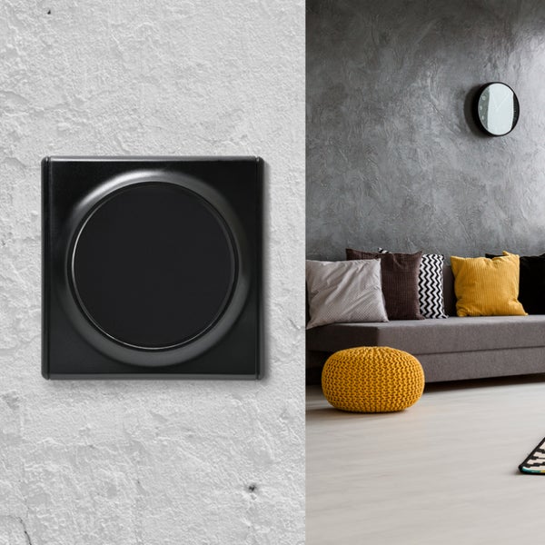 Ein moderner Lichtschalter mit rundem Einsatz auf einer strukturierten Wand neben einem Wohnraum mit Sofa und dekorativen Kissen.