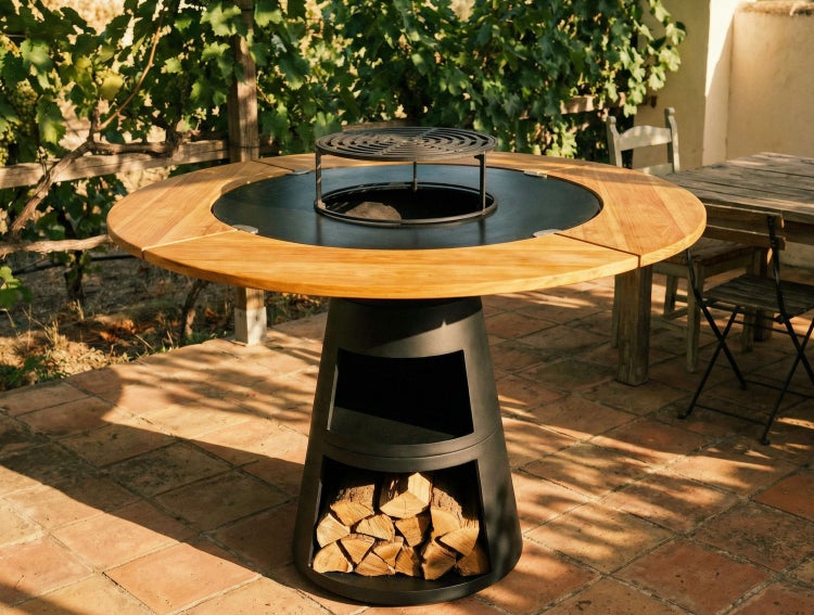 Runder Grilltisch aus Holz und schwarzem Metall mit integrierter Feuerschale, Grillrost und Holzaufbewahrung auf einer Terrasse.