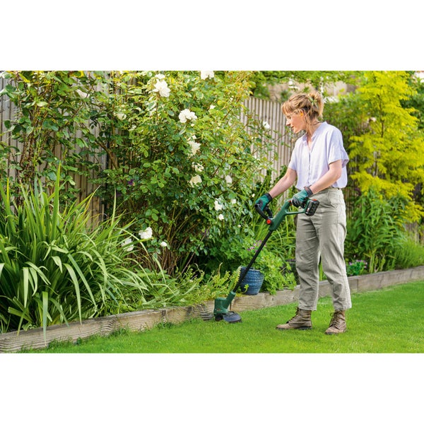 Eine Frau kürzt mit einem Akku-Rasentrimmer die Kanten eines gepflegten Rasens in einem hellen Garten mit weißen Blumen und Holzeinfassung.