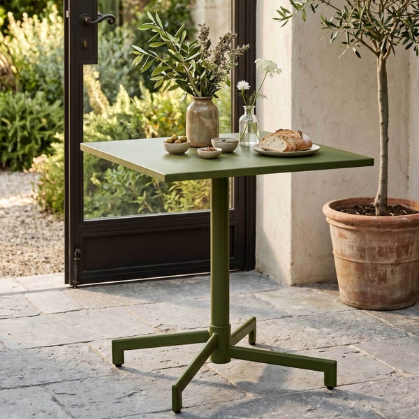 Quadratischer grüner Bistrotisch aus Metall auf einer Steinterrasse mit Blumenvasen, Schalen und Brot vor einer Gartenkulisse.