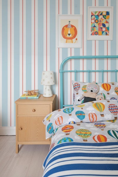 Helles Kinderzimmer mit gestreifter Tapete, blauem Metallbett, Heißluftballon-Bettwäsche und einem hölzernen Nachttisch mit Lampe.